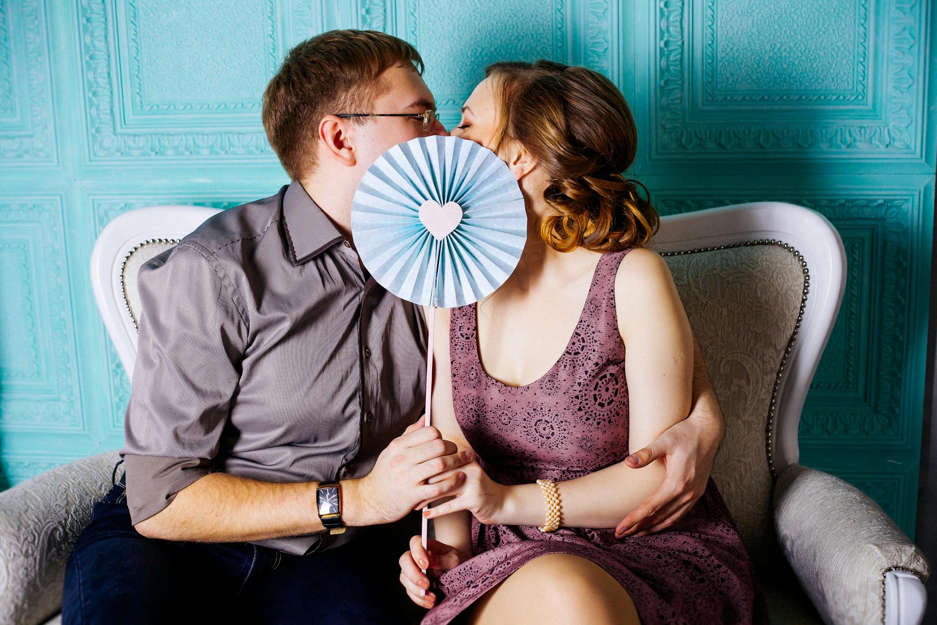 Cute white couple paired by matchmakers in Tampa sitting on a suede couch while kissing behind a blue fan