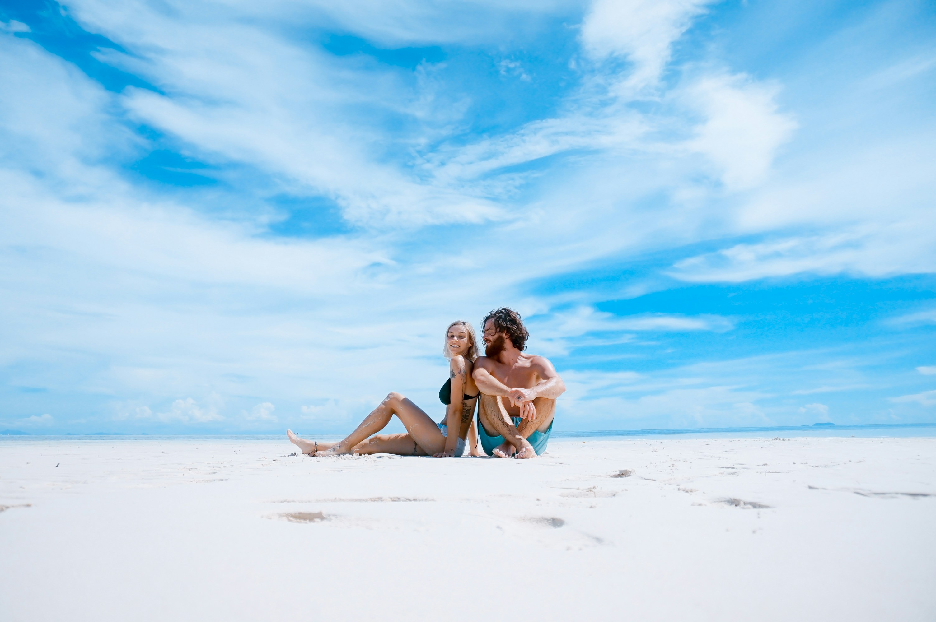 Caucasian couple sitting on a beach smiling at each other with a blue sky in the background