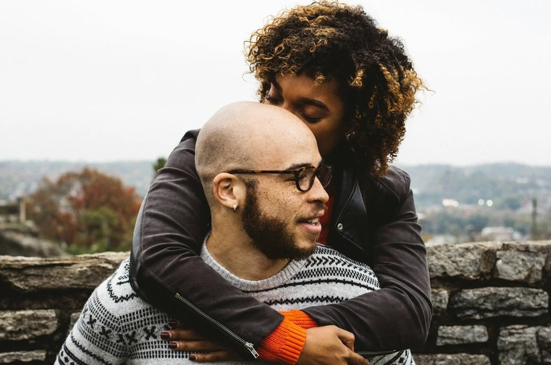 African American celibate dating couple hugging outside near a stone wall in colder weather attire